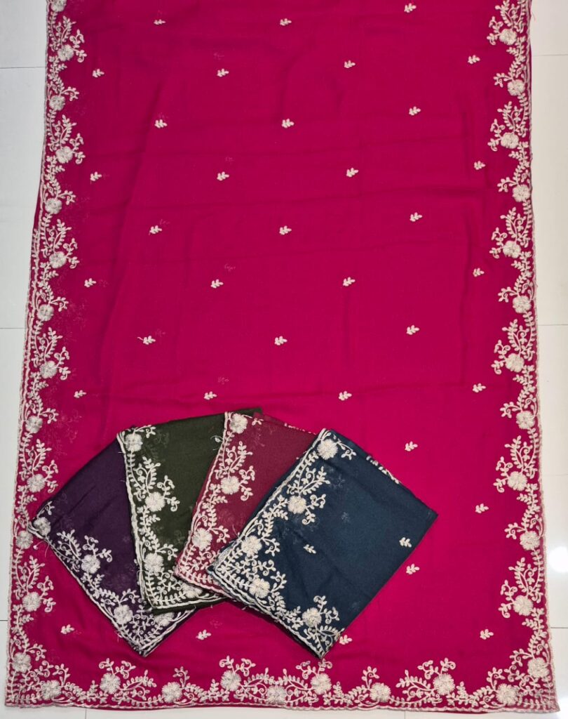 A full-frame product shot of a magenta saree with a wide floral embroidered border. Three other folded sarees in purple, olive green, and dark blue are stacked on the main saree.