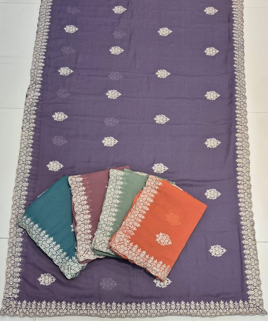 A full-frame product shot of a dark purple saree with a wide floral embroidered border. Four other folded sarees in teal, dusty rose, light green, and orange are stacked on the main saree.
