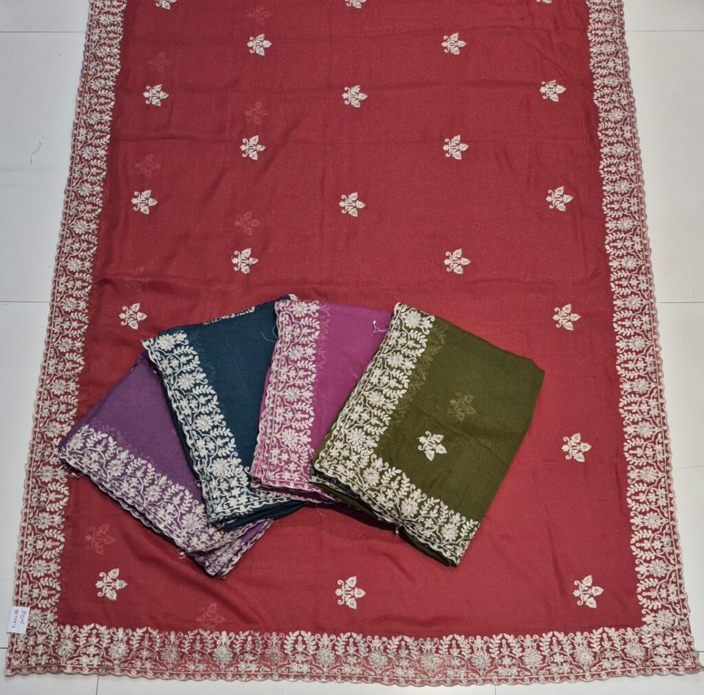A full-frame product shot of a maroon saree with a wide floral embroidered border. Three other folded sarees in purple, teal, and magenta are stacked on the main saree.