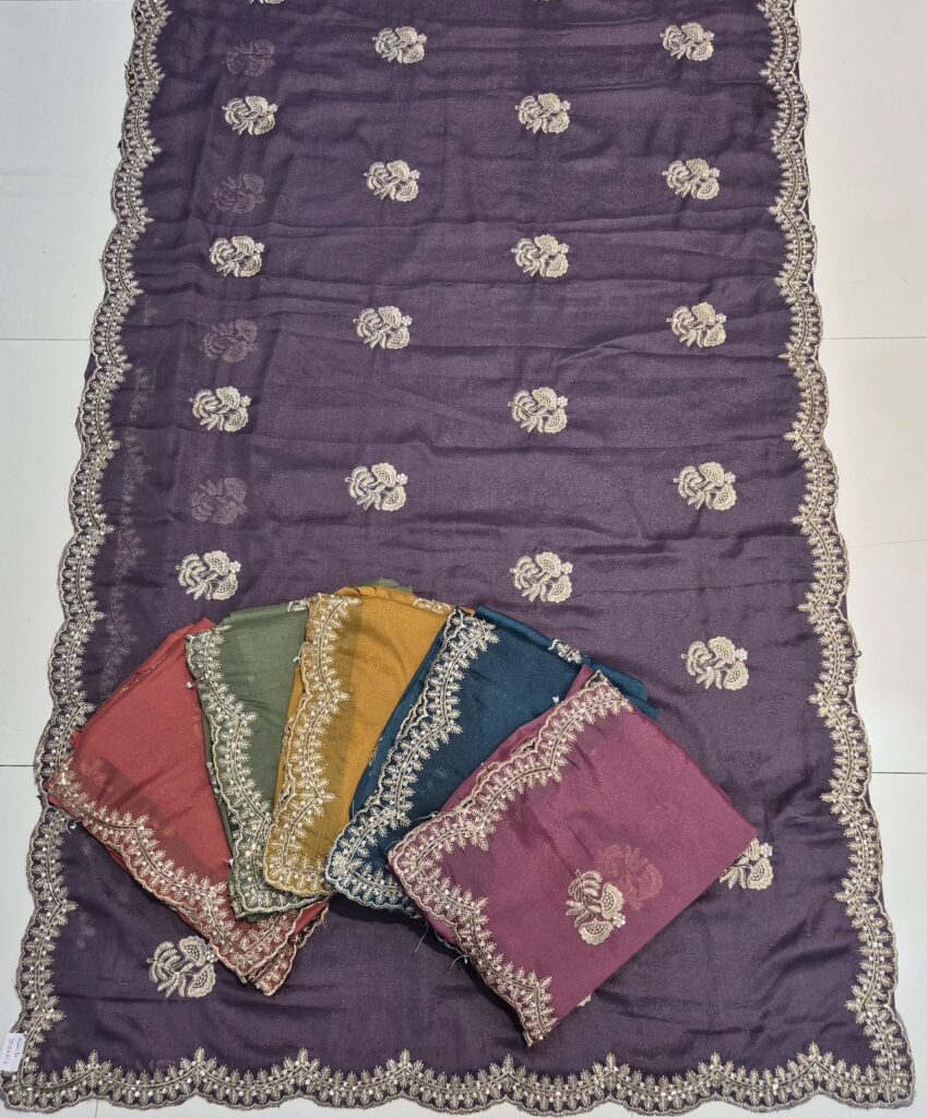 A full-frame product shot of a dark purple saree with a wide floral embroidered border. Four other folded sarees in maroon, olive green, mustard yellow, and teal are stacked on the main saree.