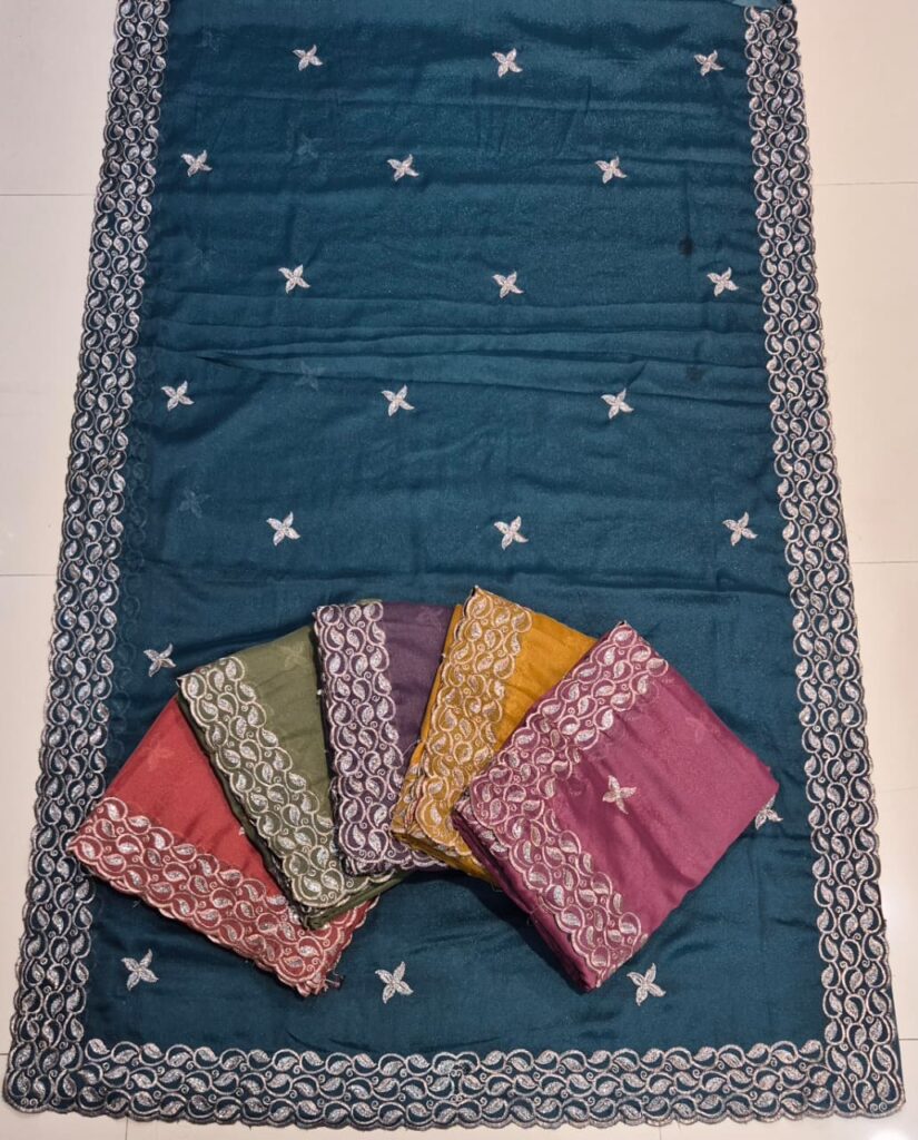 A set of colorful embroidered sarees neatly displayed on a dark teal saree with white floral embroidery and an ornate border. The sarees come in shades of pink, green, purple, mustard yellow, and magenta, each featuring a decorative lace border.