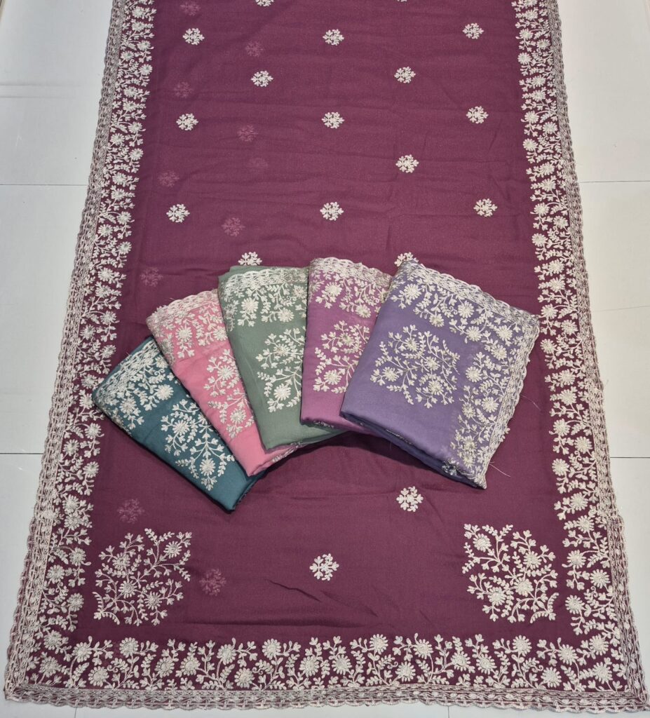 A full-frame product shot of a deep purple saree with a wide floral embroidered border. Four other folded sarees in teal, baby pink, light green, and lavender are stacked on the main saree.