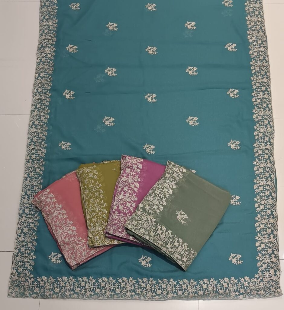 A full-frame product shot of a teal saree with a wide floral embroidered border. Three other folded sarees in pink, green, and purple are stacked on the main saree.