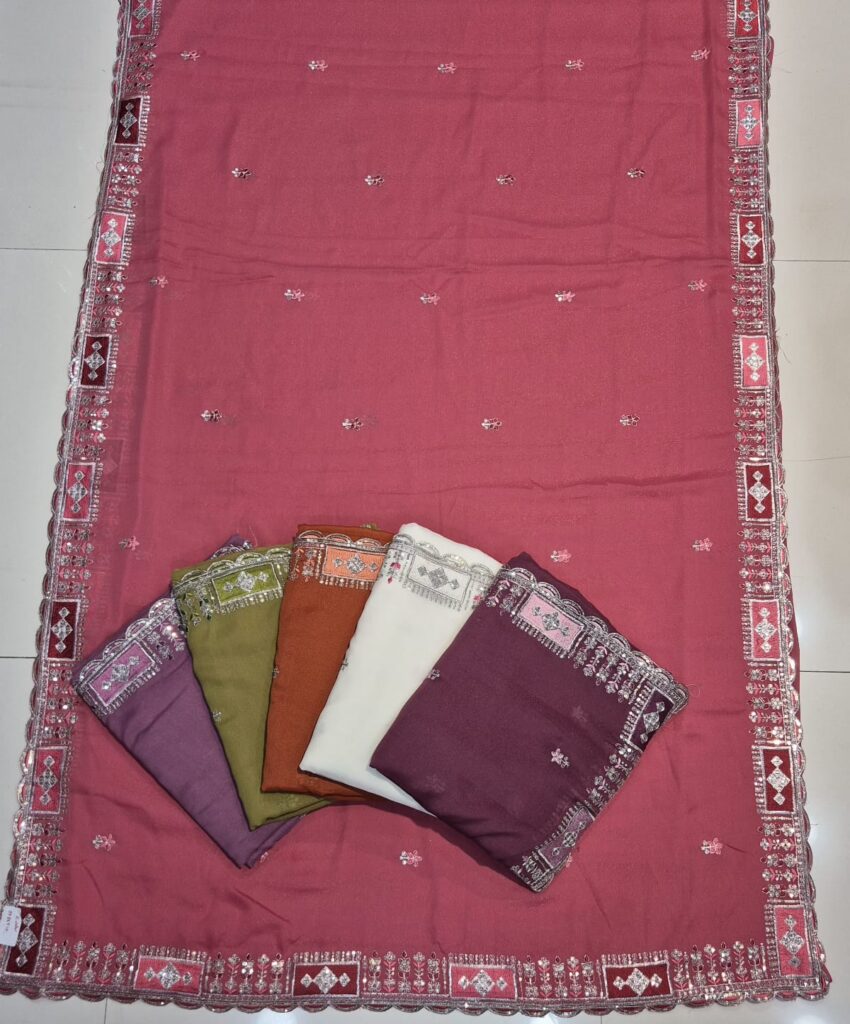 A full-frame product shot of a dusty rose saree with a traditional embroidered border and scattered sequin work. Four folded sarees in purple, light green, rust orange, and off-white are stacked on the main saree.