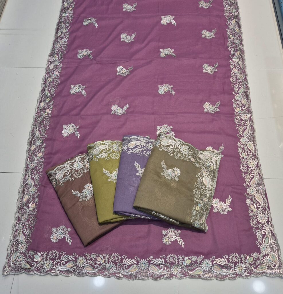 A full-frame product shot of a purple saree with a paisley- and floral-embroidered border. Three other sarees in brown, light green, and purple are folded and placed on top of the main saree, arranged in a row from left to right. The main saree has a delicate embroidery pattern scattered throughout the body. The sarees are displayed on a white tiled floor.